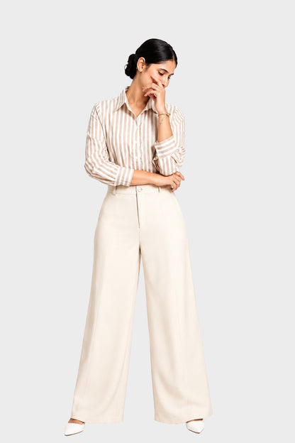 A woman modeling a cream and beige vertically striped button-down shirt paired with high-waisted, wide-leg cream trousers, set against a solid light gray background.