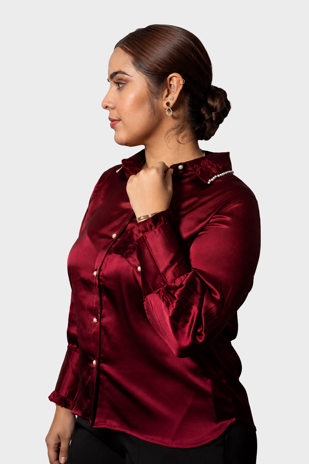 A woman in profile wearing a shiny burgundy satin button-up blouse with ruffled details, photographed against a plain light gray studio background.