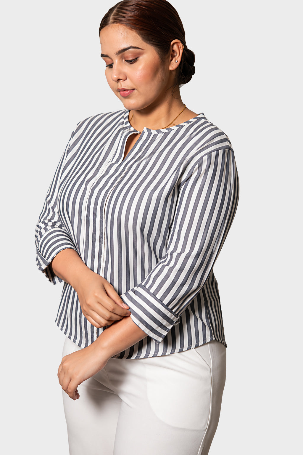 A woman wearing a navy and white vertically striped long-sleeve blouse with a split neckline, paired with white pants, photographed against a light gray studio background.