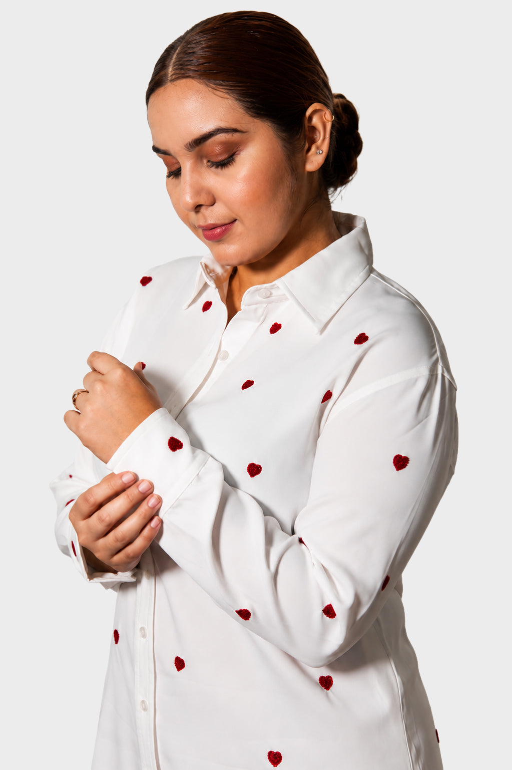 A woman wearing a white long-sleeve button-up shirt with small red heart embroidery, shown from the chest up against a light gray studio background.