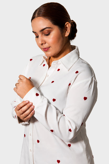 A woman wearing a white long-sleeve button-up shirt with small red heart embroidery, shown from the chest up against a light gray studio background.