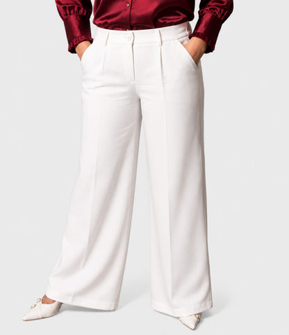 A model wearing white wide-leg trousers with pleats, paired with a dark red satin shirt and white pointed-toe flats, shown from the waist down on a light gray background.