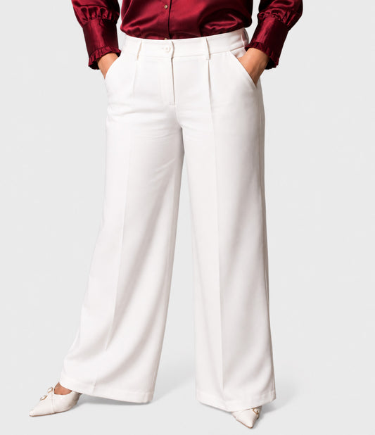 A model wearing white wide-leg trousers with pleats, paired with a dark red satin shirt and white pointed-toe flats, shown from the waist down on a light gray background.