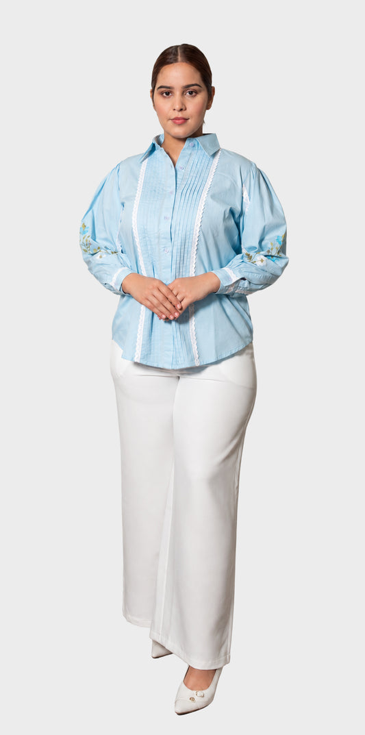 A woman modeling a light blue long-sleeve button-up shirt with white vertical stripes and embroidered details on the sleeves, paired with white wide-leg pants and white pointed shoes, against a light gray background.