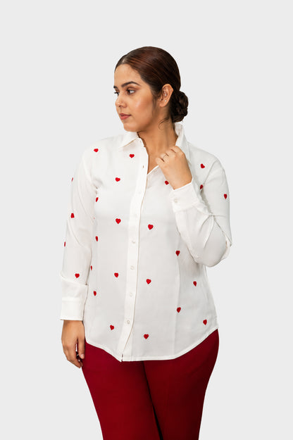 A woman modeling a white long-sleeve button-up shirt with small red heart patterns, paired with dark red pants, against a solid light gray background.