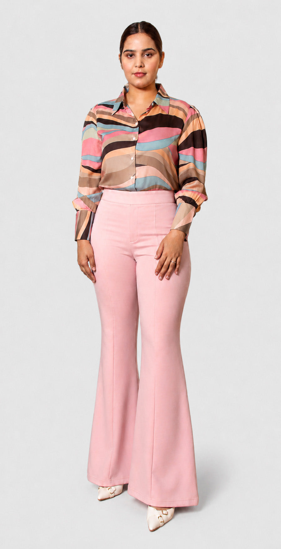 A woman modeling a long-sleeve button-up shirt with horizontal multicolor stripes in pink, beige, brown, and blue, paired with high-waisted light pink flared pants, against a light gray studio background.