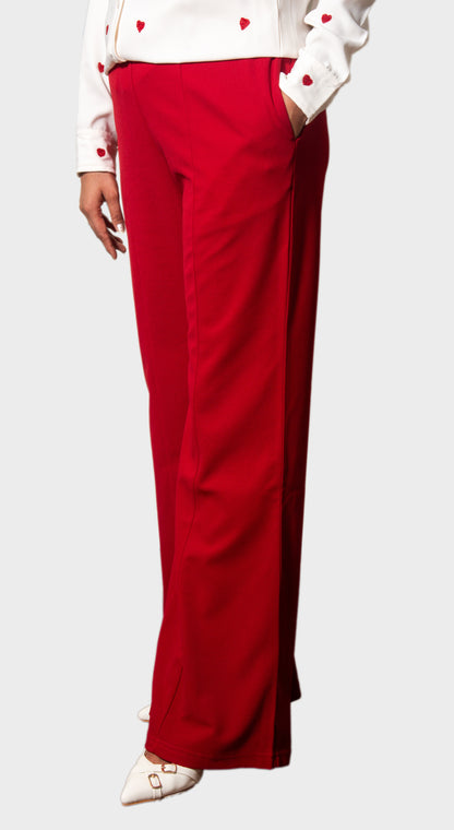 A person wearing bright red wide-leg trousers with a white long-sleeve top and white pointed shoes, shown from the chest down against a light gray studio background.