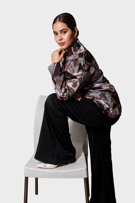 A woman poses on a white chair wearing a long-sleeve high-neck blouse with a brown, black, and beige abstract pattern, paired with black wide-leg trousers and white flat shoes, against a light gray studio background.