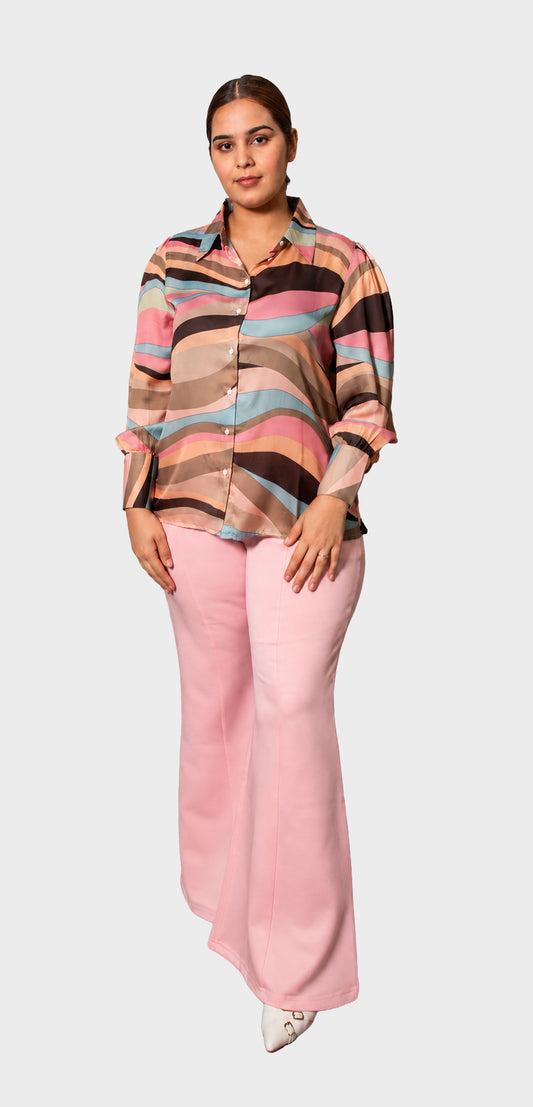 A woman modeling a long-sleeve button-up shirt with wavy horizontal stripes in pink, beige, brown, and light blue, paired with light pink wide-leg pants, against a light gray background.