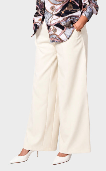 A person wearing off-white wide-leg trousers with a patterned top and white pointed-toe heels, shown from the chest down against a light gray studio background.