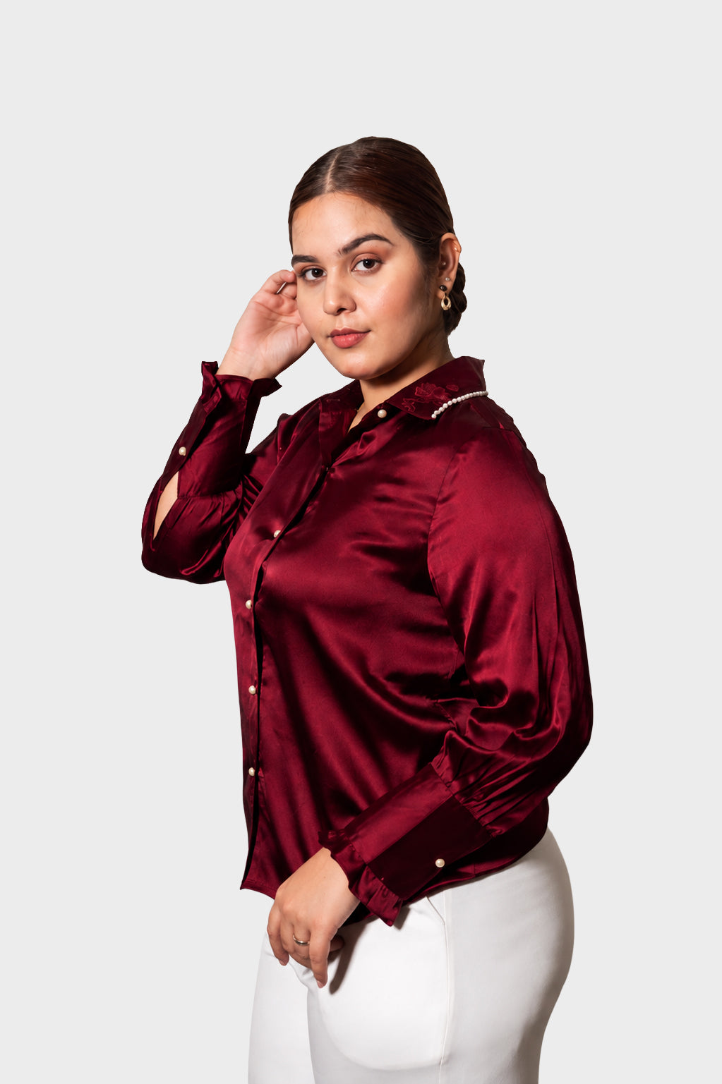 A woman modeling a long-sleeve satin blouse in deep burgundy with gold buttons, paired with white pants, against a solid light gray background.