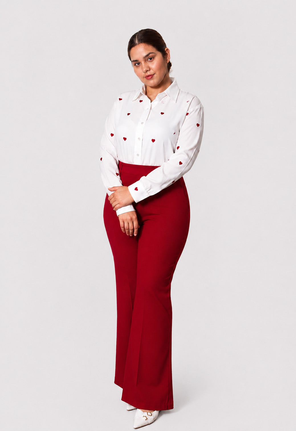 A woman modeling a white long-sleeve button-up shirt with small red heart details and high-waisted, wide-leg red pants against a light gray studio background.