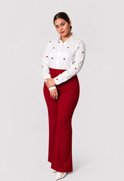 A woman modeling a white long-sleeve button-up shirt with small red heart details and high-waisted, wide-leg red pants against a light gray studio background.