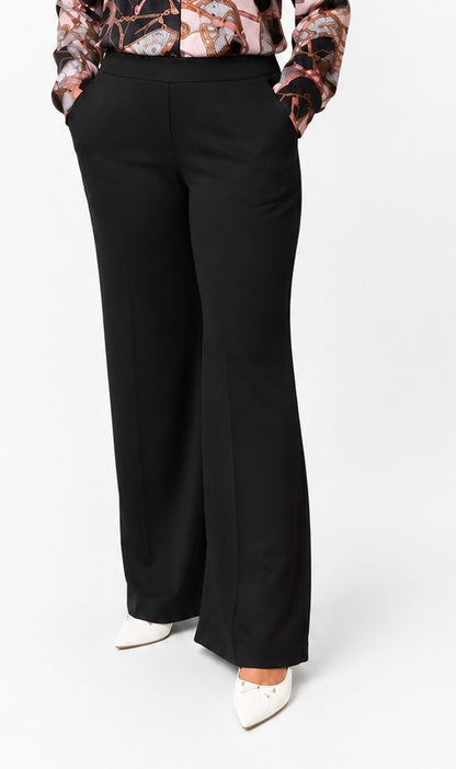 A model wearing black wide-leg trousers, paired with a patterned blouse and white pointed-toe heels, shown from the waist down against a plain white studio background.