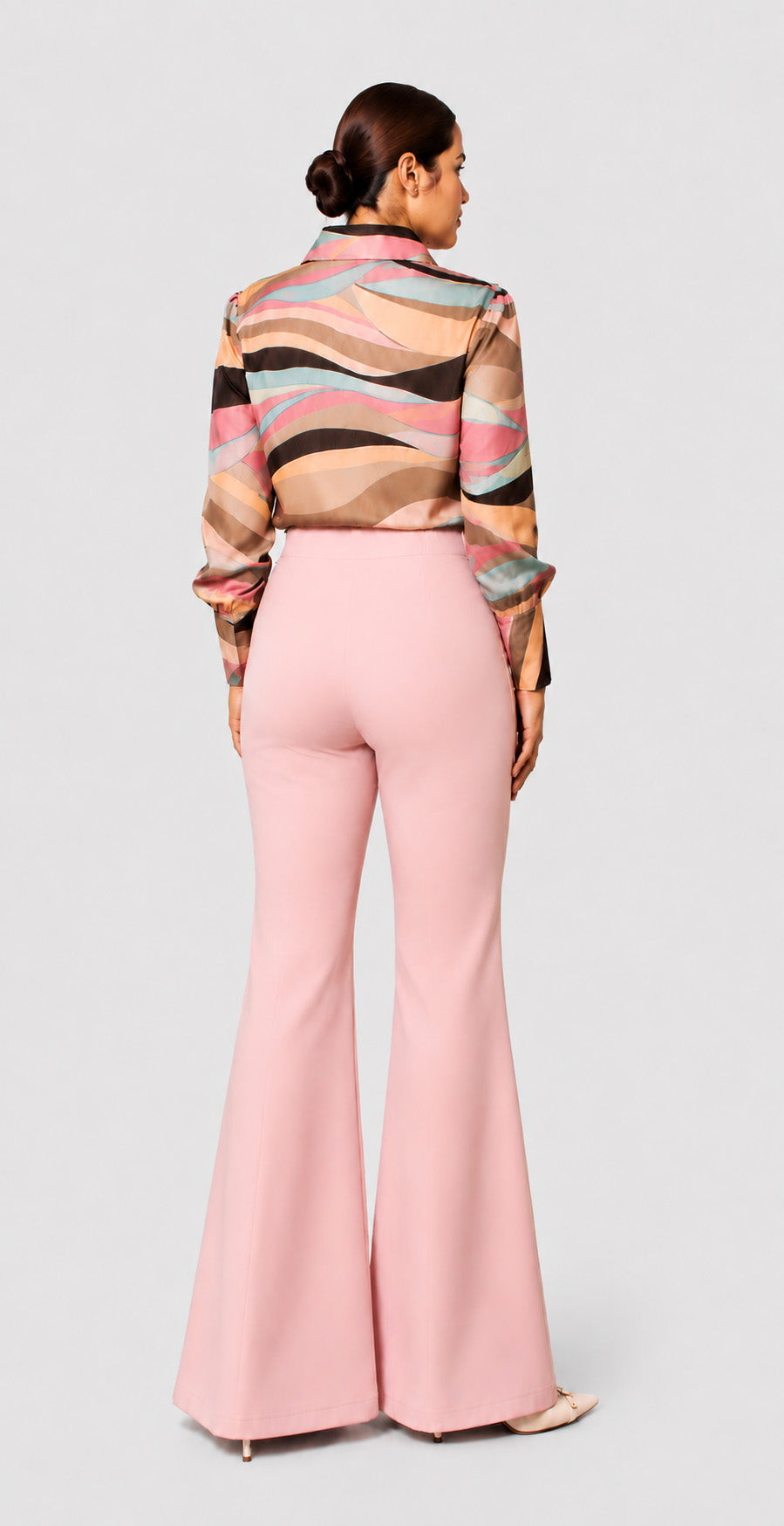 A woman shown from the back wearing a long-sleeve collared blouse with a wavy multicolor pattern in beige, brown, black, and pink, paired with light pink flared pants, against a light gray studio background.