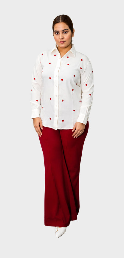 A woman modeling a white long-sleeve button-up shirt with small red embroidered motifs and dark red wide-leg pants, standing against a light gray studio background.