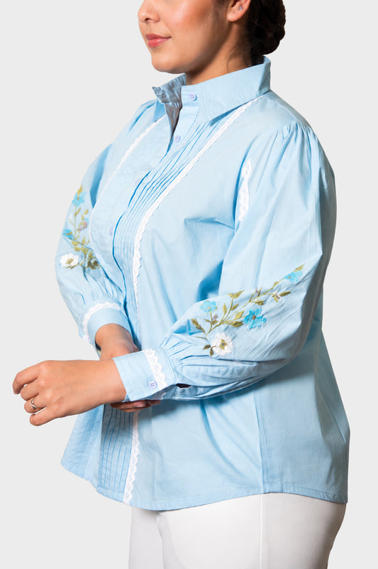 A woman wearing a light blue long-sleeve button-up blouse with white lace trim and floral embroidery on the sleeves, paired with white pants, against a light gray studio background.