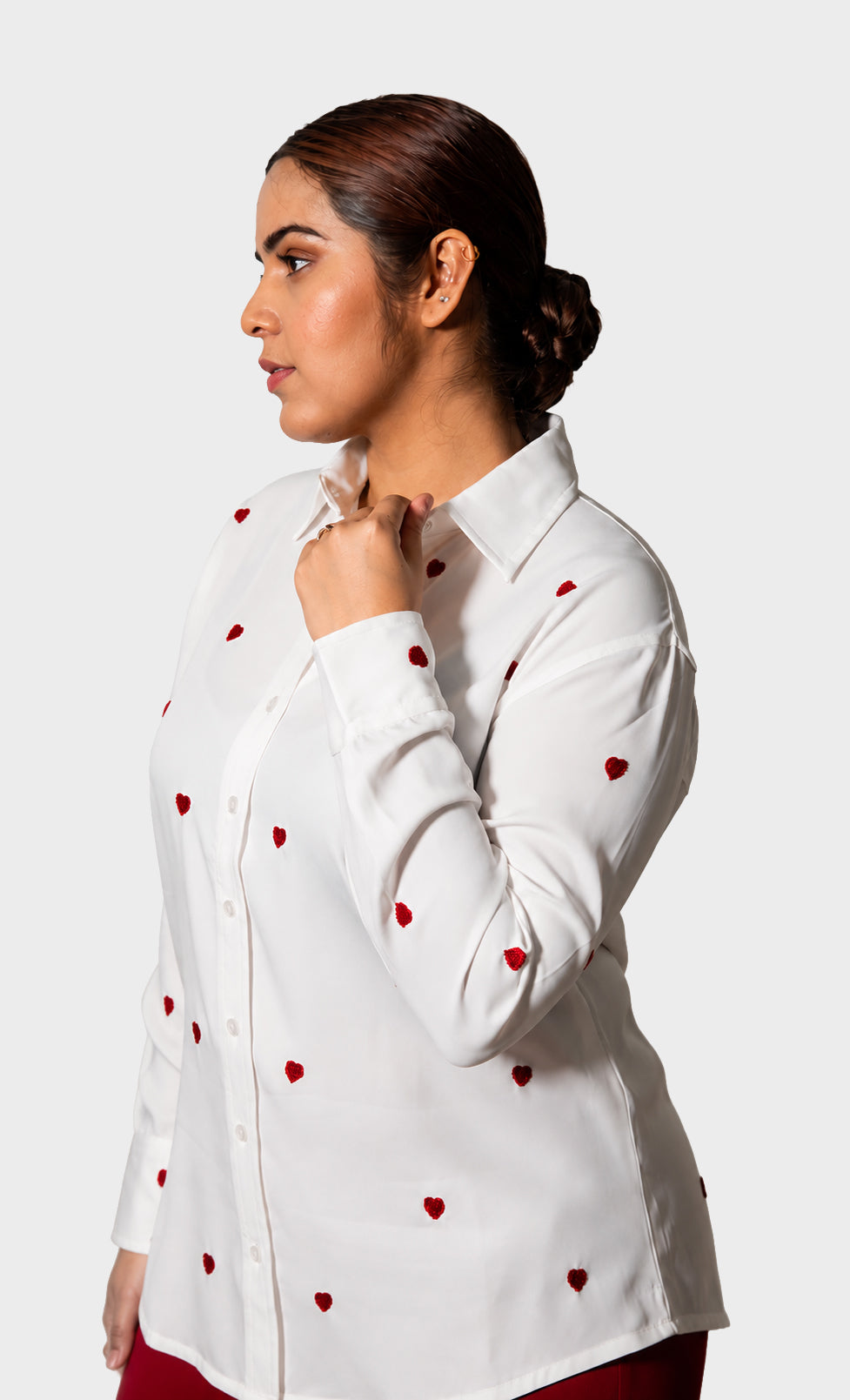 A woman in profile wearing a white long-sleeve button-up shirt with small red heart motifs, posed against a light gray studio background.