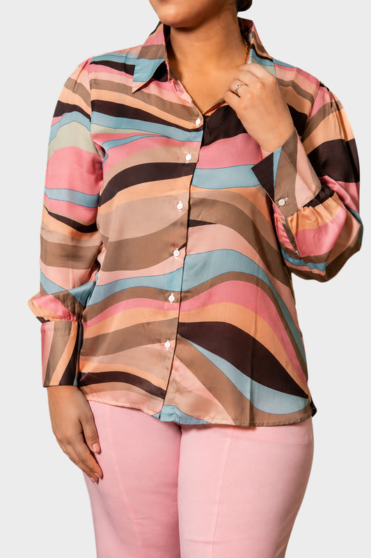 A woman wearing a long-sleeve button-up blouse with a wavy abstract stripe pattern in pink, beige, brown, and light blue, paired with pink pants, against a light gray background.
