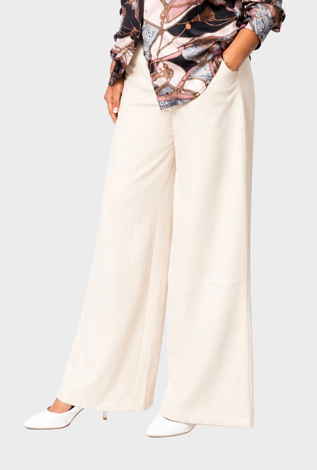 A model wearing high-waisted, wide-leg cream trousers paired with a patterned blouse and white pointed-toe heels, shown from the waist down against a light gray studio background.