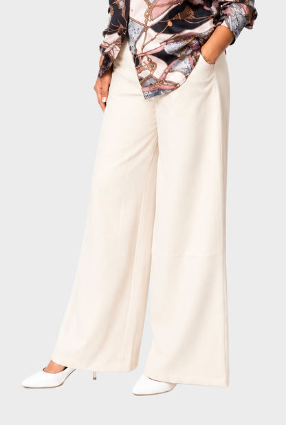A model wearing high-waisted, wide-leg cream trousers paired with a patterned blouse and white pointed-toe heels, shown from the waist down against a light gray studio background.