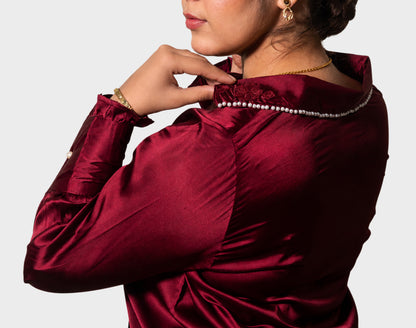 A close-up of a woman wearing a deep burgundy satin blouse with a high collar decorated with a line of white pearls, shown from a side-back angle against a light gray background.