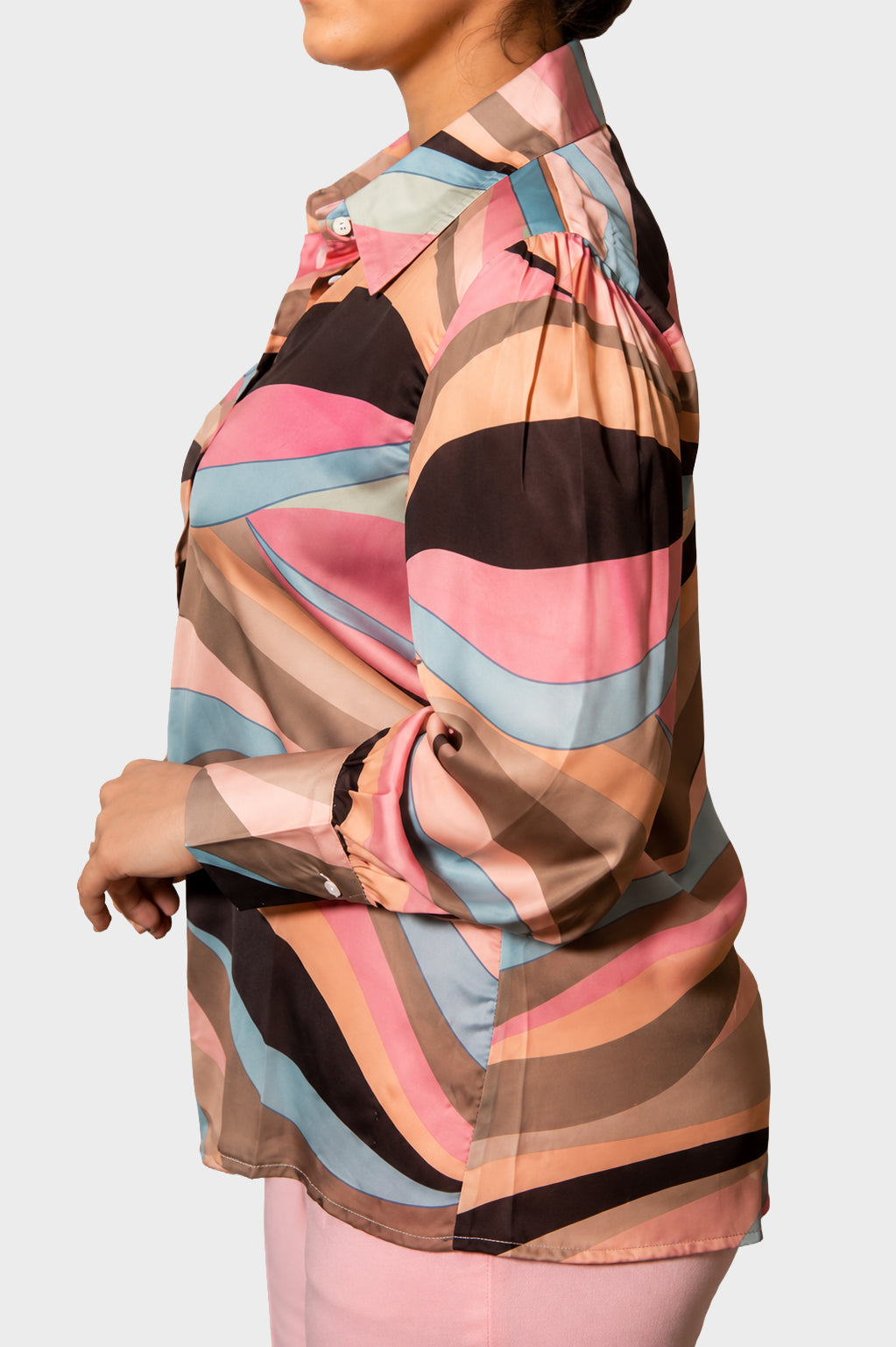A woman shown from the side wearing a long-sleeve button-up blouse with a wavy abstract stripe pattern in pink, beige, brown, light blue, and black, paired with light pink pants against a light gray studio background.