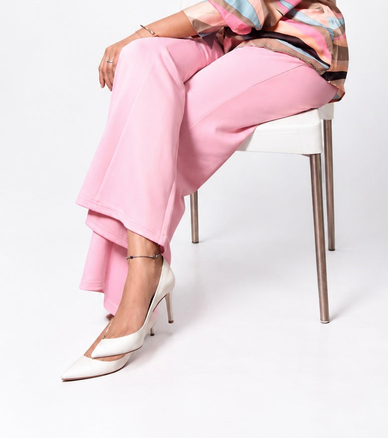 A model seated on a white chair wearing pink wide-leg trousers, a multicolor patterned top, and white pointed-toe slingback heels against a clean white studio background.