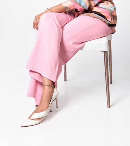 A model seated on a white chair wearing pink wide-leg trousers, a multicolor patterned top, and white pointed-toe slingback heels against a clean white studio background.