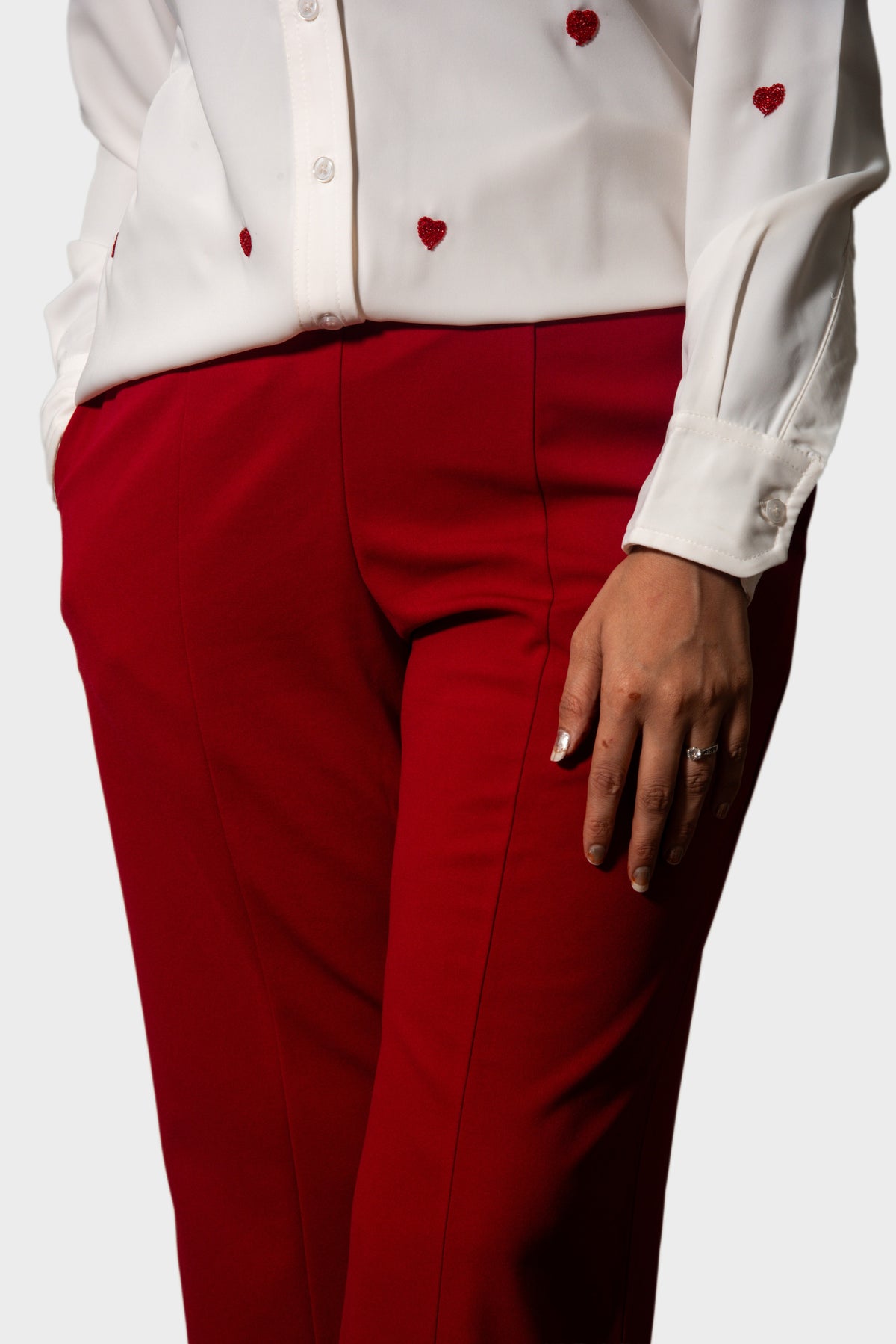 A close-up of a person wearing bright red tailored trousers and a white button-up blouse with small red heart motifs, photographed against a light gray studio background.
