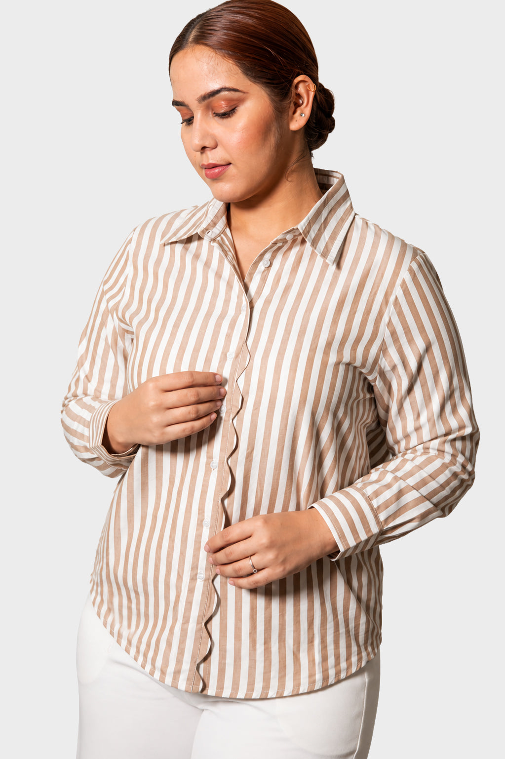 A woman wearing a beige and white vertically striped button-up shirt with scalloped edges, paired with white pants, against a light gray studio background.