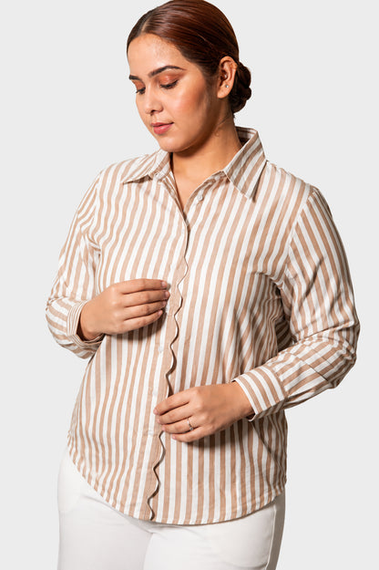 A woman wearing a beige and white vertically striped button-up shirt with scalloped edges, paired with white pants, against a light gray studio background.