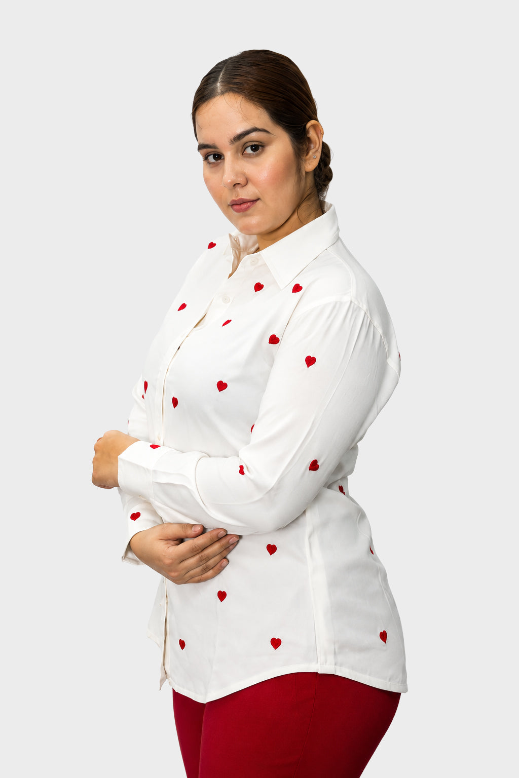 A woman modeling a white long-sleeve button-up shirt with small red heart prints, paired with red pants, against a light gray studio background.