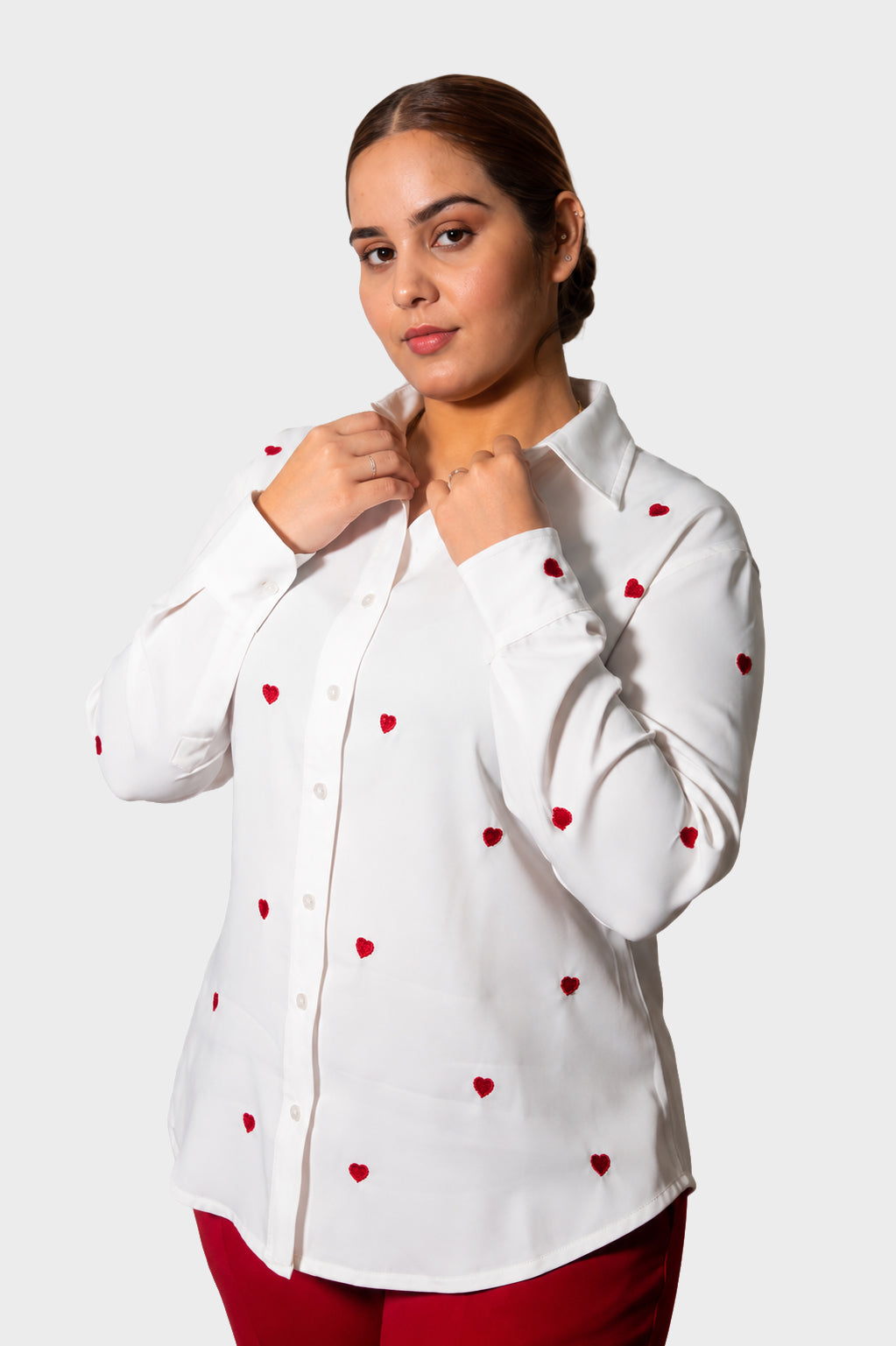 A woman wearing a white long-sleeve button-up shirt with small red heart embroidery, posing against a light gray background.