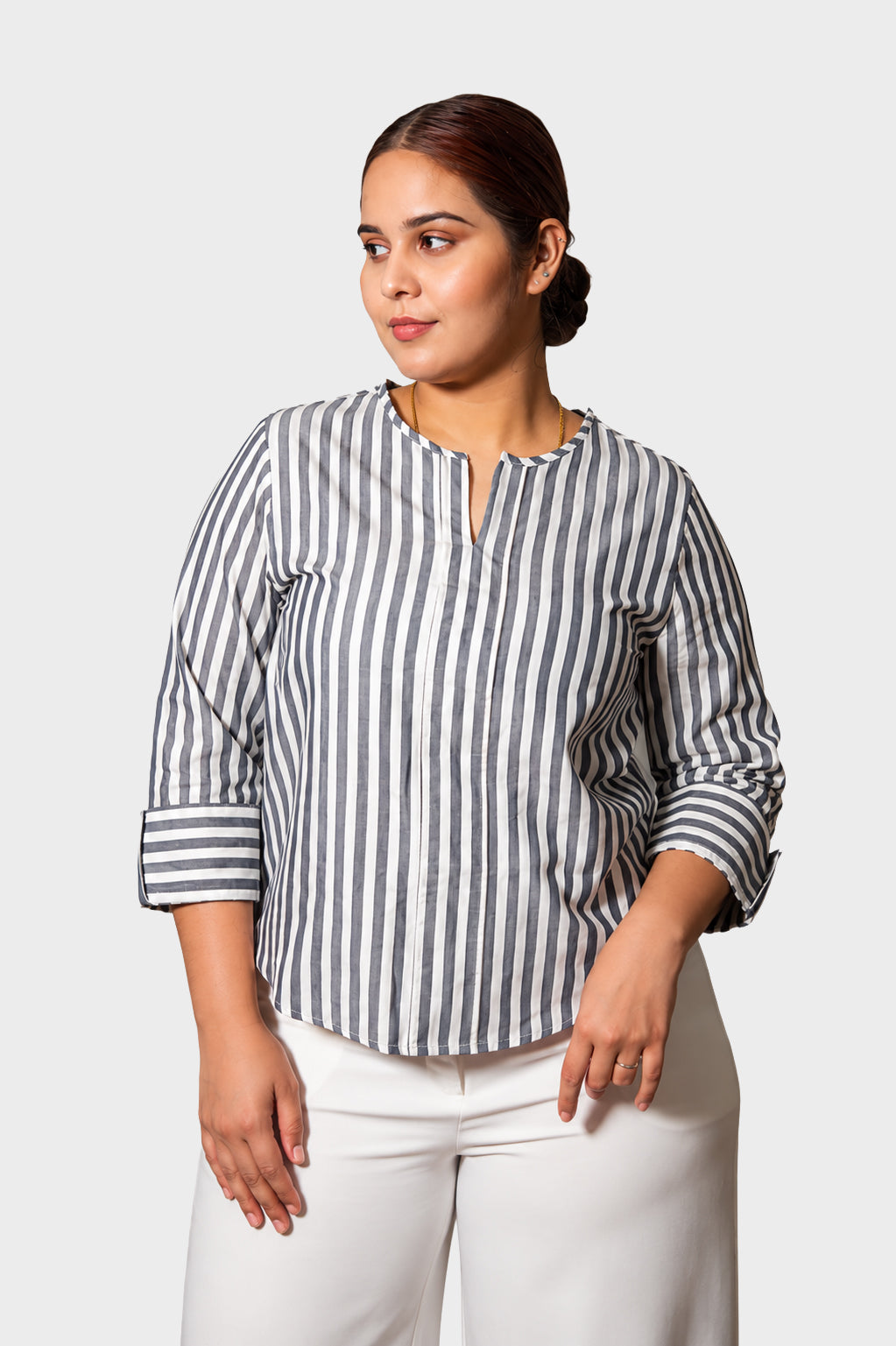 A woman modeling a navy and white vertically striped long-sleeve blouse with a split neckline, paired with white pants, against a light gray studio background.