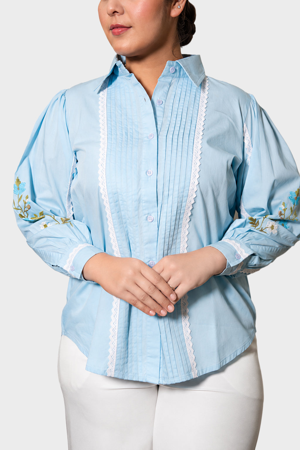 A woman wearing a light blue Guayabera-style blouse with white lace trim down the front and floral embroidery on the sleeves, paired with white pants against a light gray studio background.