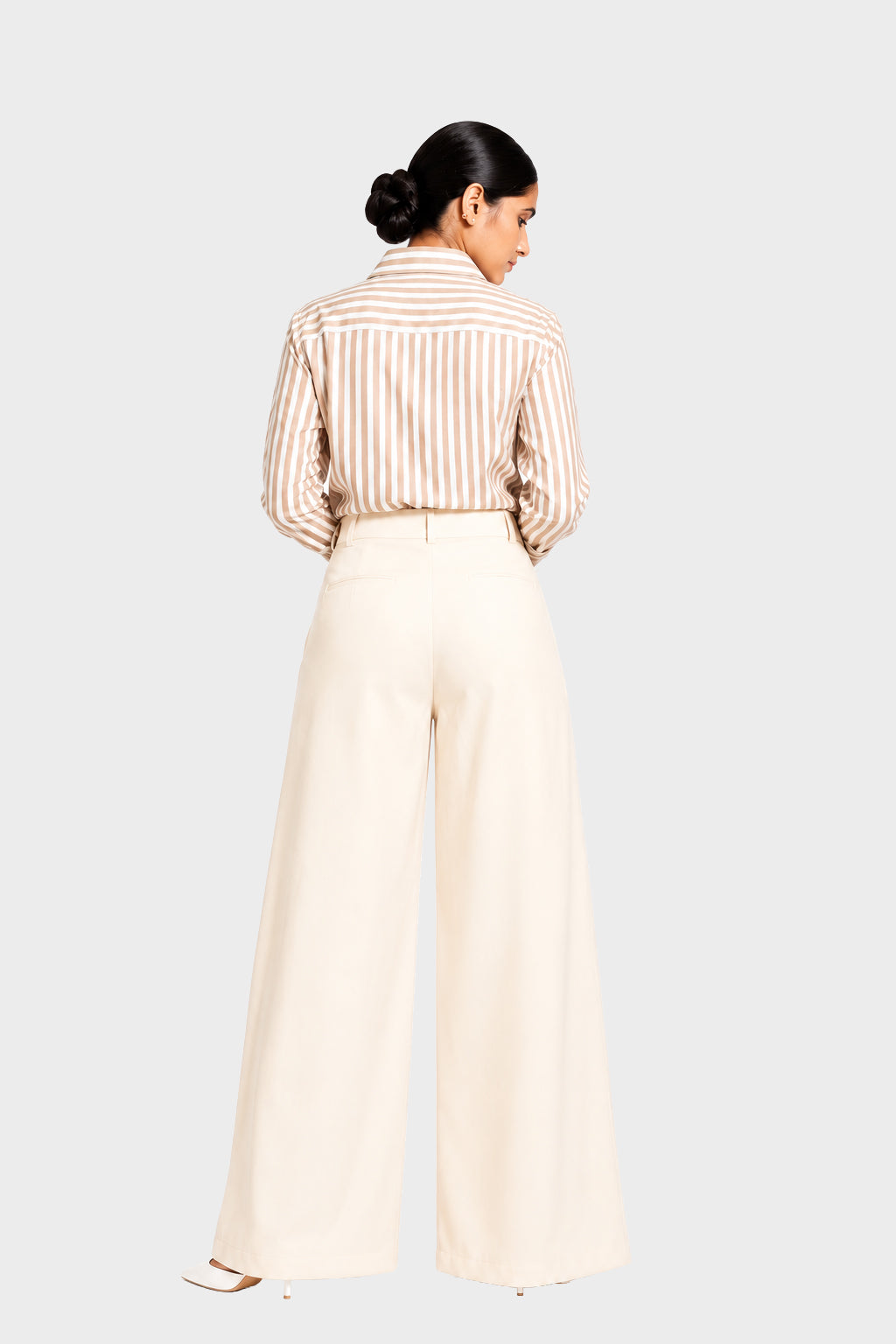 A woman shown from the back wearing a beige and white vertically striped button-down shirt tucked into high-waisted, wide-leg cream trousers, set against a solid light gray background.