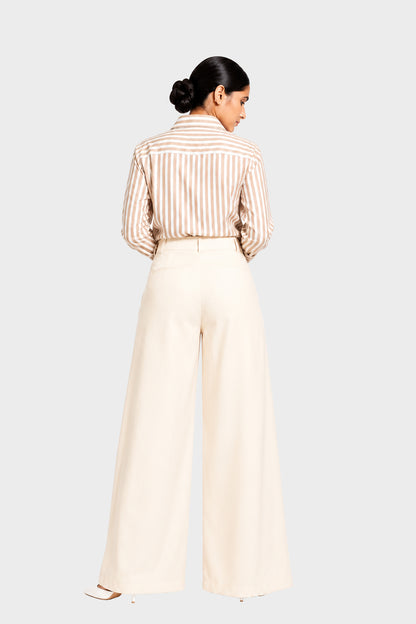 A woman shown from the back wearing a beige and white vertically striped button-down shirt tucked into high-waisted, wide-leg cream trousers, set against a solid light gray background.