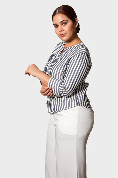 A woman modeling a black and white vertically striped long-sleeve blouse with a split neckline, paired with white trousers, against a light gray studio background.