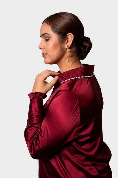 A woman in profile wearing a shiny deep burgundy satin blouse with a pearl-trimmed collar, photographed against a light gray studio background.