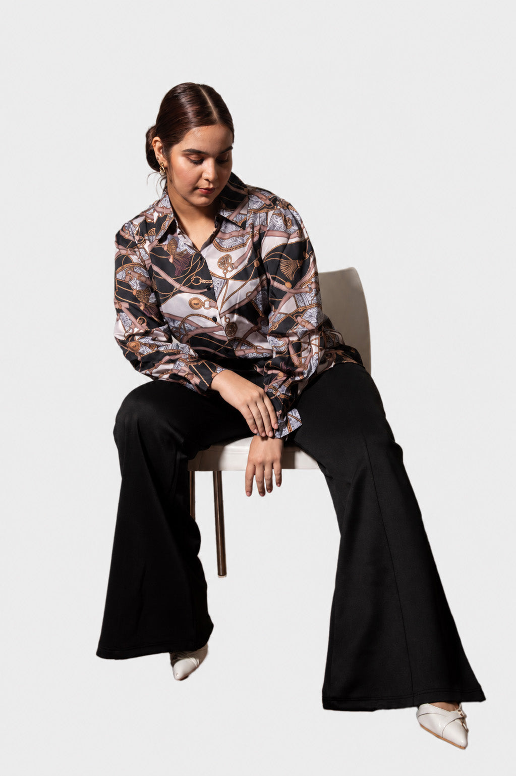 A woman sits on a minimalist chair against a light gray background, wearing a long-sleeve button-up blouse with a brown, beige, and black abstract pattern, paired with black wide-leg pants and white pointed shoes.