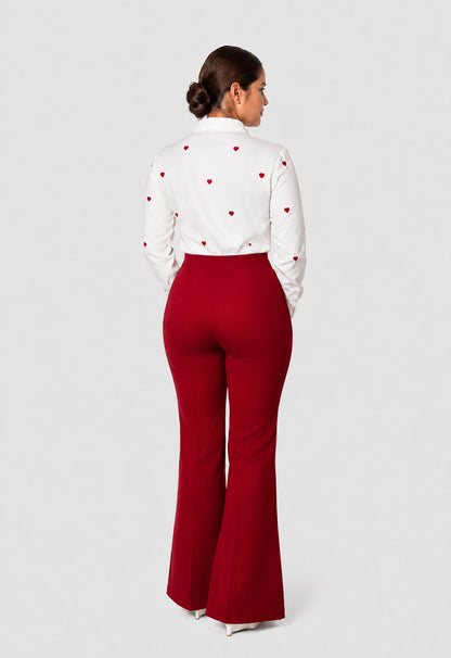 A woman shown from the back wearing a white long-sleeve blouse with small red heart motifs and high-waisted red flared pants, photographed against a light gray studio background.