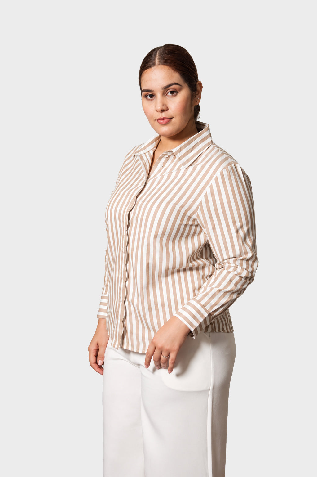 A woman modeling a beige and white vertically striped button-down shirt with white pants, photographed against a solid light gray studio background.