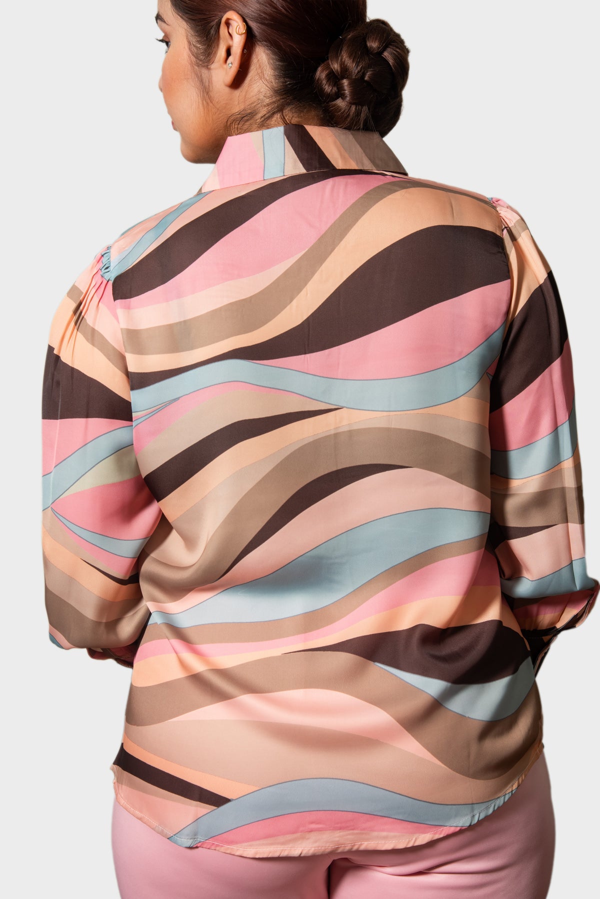A woman shown from the back wearing a long-sleeve collared blouse with a wavy abstract stripe pattern in pink, beige, brown, and light blue, paired with pink pants against a light gray studio background.