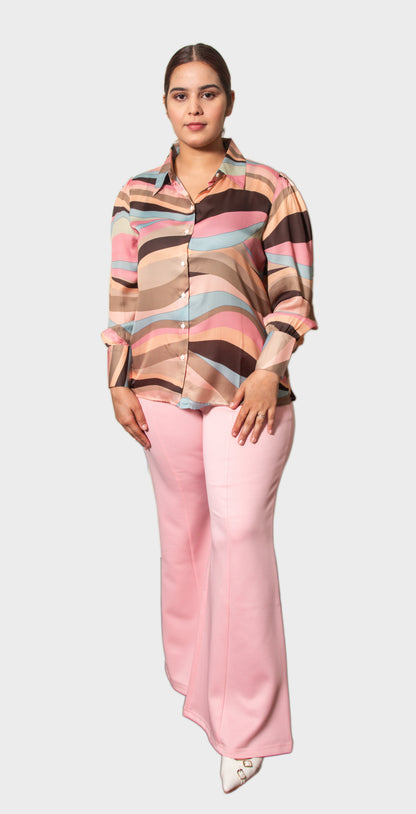 A woman modeling a long-sleeve button-up shirt with a wavy multicolor stripe pattern in pink, beige, brown, and light blue, paired with light pink wide-leg pants, against a solid light gray background.