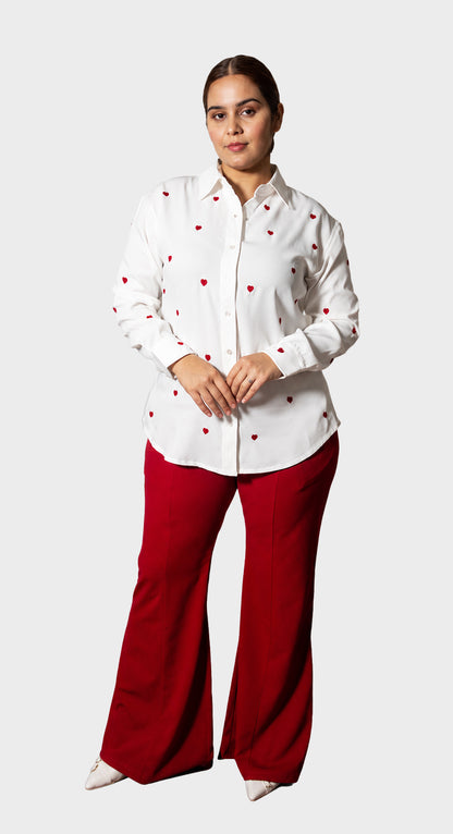A woman modeling a white long-sleeve button-up shirt with small red heart prints and bright red wide-leg pants, standing against a light gray studio background.