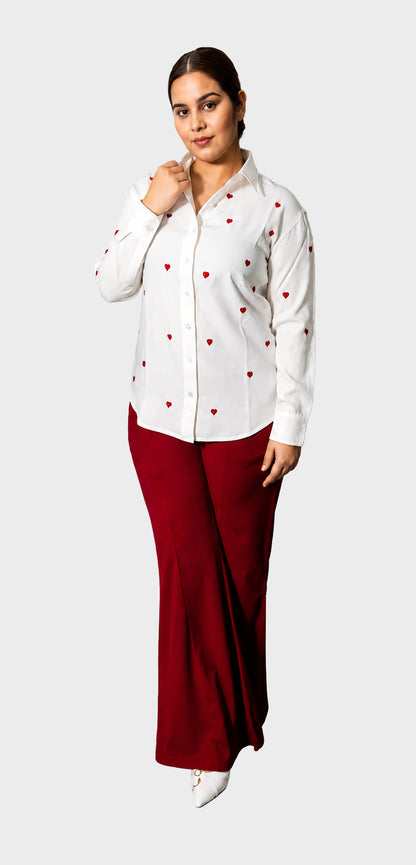 A woman modeling a white long-sleeve button-up shirt with small red heart prints, paired with dark red wide-leg pants, against a light gray background.