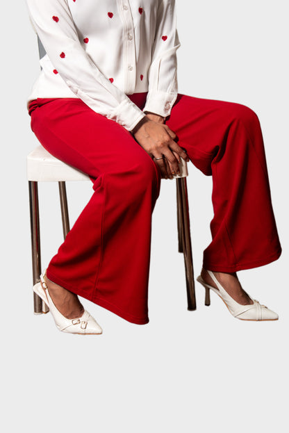 A model seated on a white stool wearing bright red wide-leg pants, a white long-sleeve top with small red heart details, and white pointed-toe heels against a light gray background.