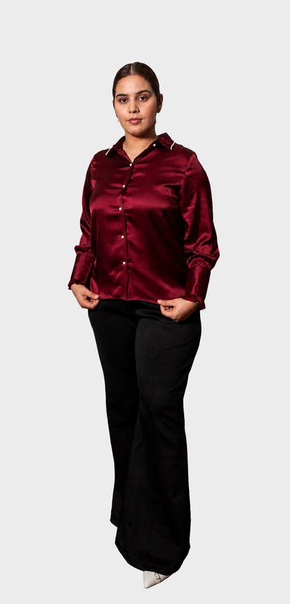 A woman modeling a long-sleeve satin button-up shirt in deep burgundy, paired with black pants, against a solid light gray background.