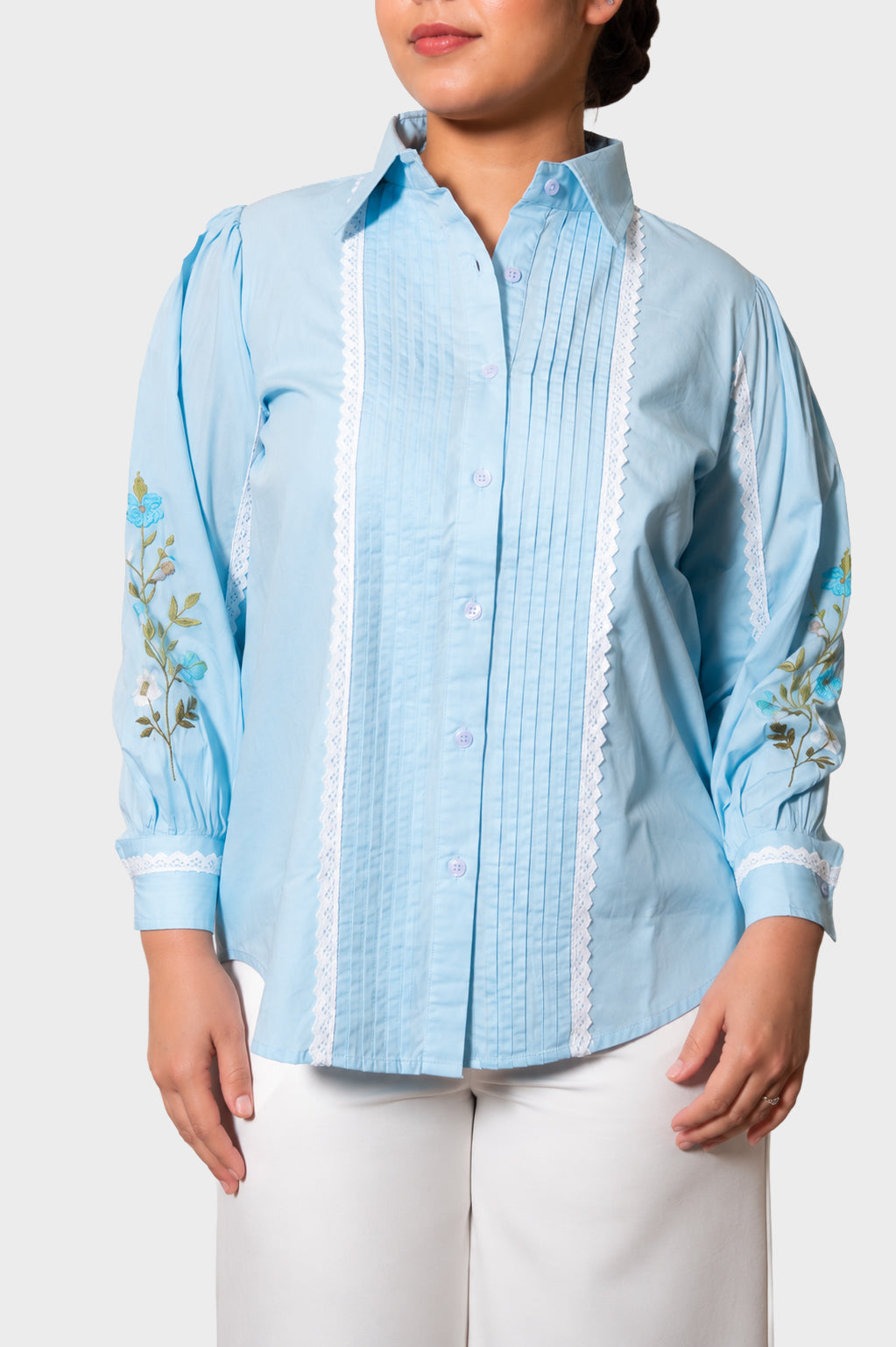 A woman wearing a light blue guayabera-style blouse with white lace trim and floral embroidery on the sleeves, paired with white pants, against a light gray studio background.